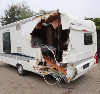 Ein beschädigter Wohnwagen mit einem großen Loch auf der Rückseite, geparkt auf einem gepflasterten Platz.